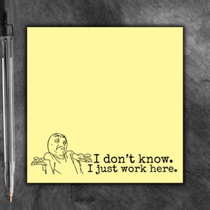 May include: A square, pale yellow sticky note featuring a black line-art illustration of a person shrugging with the text "I don't know. I just work here." positioned next to a black ballpoint pen with a clear plastic barrel and metal clip.