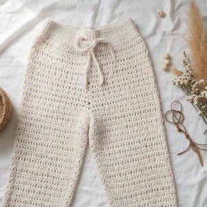 May include: Cream-colored crochet pants with a drawstring waist. The pants are laid flat on a white sheet, with a woven basket and a bouquet of dried flowers and grasses nearby. The crochet stitch creates a textured look.