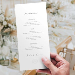 May include: A white menu card with elegant script text, held in a hand, against a blurred background of a wedding reception. The menu lists starters, mains, and dessert. The names "Julia & Adrian" are at the bottom.