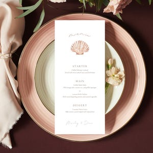 May include: A formal table setting with a menu card. The menu card features a seashell graphic and text. The plate is pink with a gold rim, and the silverware is gold. A folded napkin and flowers complete the elegant arrangement.