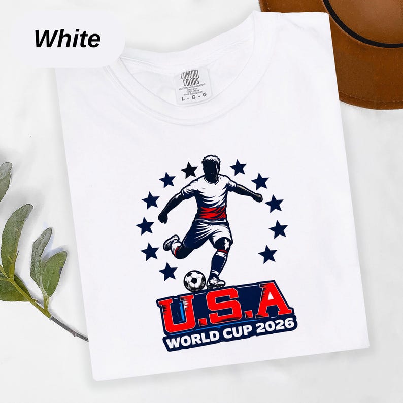 Retro USA World Cup Shirt 2026 design featuring a silhouette of a soccer player kicking a football, surrounded by a semi-circle of blue stars with bold red and blue U.S.A. United States Soccer fan apparel typography, as shown in image