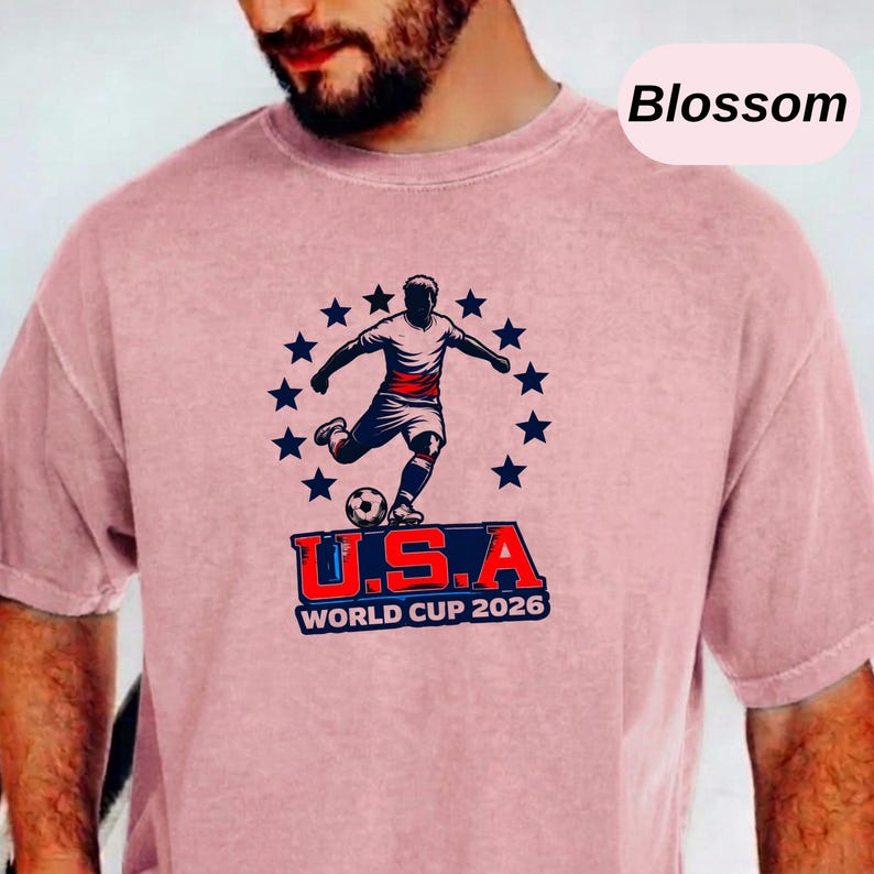 May include: A dusty rose short-sleeve t-shirt featuring a graphic of a soccer player kicking a ball surrounded by a circle of dark blue stars. Below the player, the text reads U.S.A. WORLD CUP 2026 in bold red and dark blue lettering.