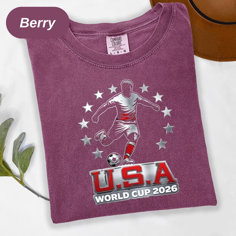 May include: A berry-colored t-shirt featuring a graphic of a soccer player kicking a ball surrounded by a circle of silver stars. Below the player, the text reads U.S.A WORLD CUP 2026 in red and silver lettering. The shirt has a crew neckline.