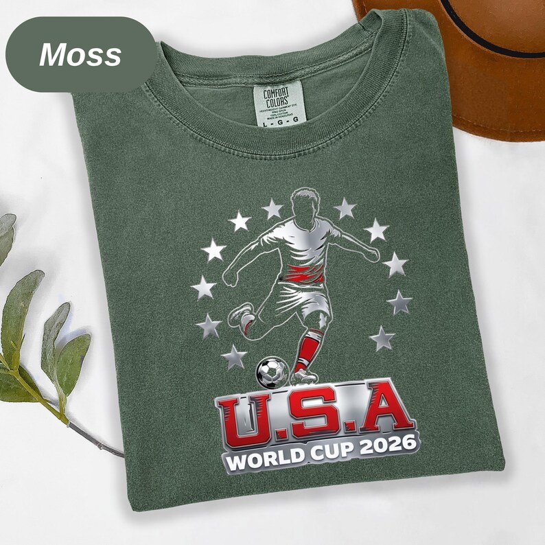May include: A moss green crew neck t-shirt featuring a graphic of a soccer player kicking a ball surrounded by a circle of white stars. Below the player, the text reads U.S.A WORLD CUP 2026 in bold red and white lettering with a metallic finish.
