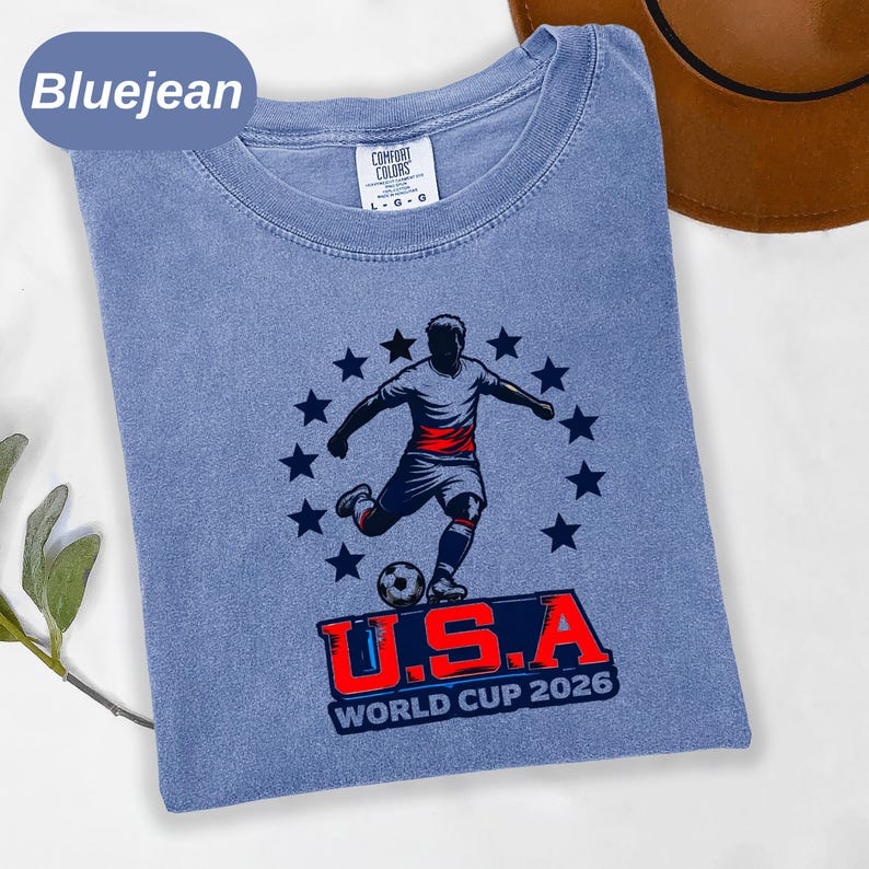 May include: A bluejean colored t-shirt featuring a graphic of a soccer player kicking a ball surrounded by a circle of dark blue stars. Below the player, the text reads U.S.A WORLD CUP 2026 in bold red and blue lettering on a dark blue background.