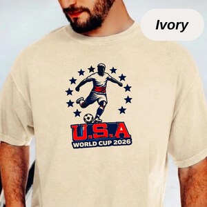 May include: An ivory short-sleeve t-shirt featuring a graphic of a soccer player kicking a ball, surrounded by a circle of dark blue stars. Below the player, the text reads U.S.A WORLD CUP 2026 in bold red, white, and blue lettering.