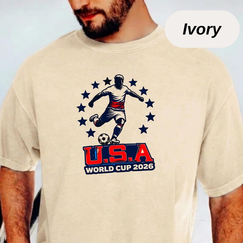 May include: An ivory short-sleeve t-shirt featuring a graphic of a soccer player kicking a ball, surrounded by a circle of dark blue stars. Below the player, the text reads U.S.A WORLD CUP 2026 in bold red, white, and blue lettering.