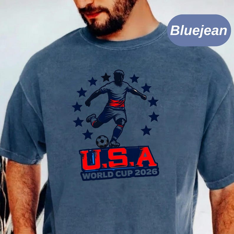 May include: A blue jean colored t-shirt featuring a graphic of a soccer player kicking a ball surrounded by a circle of stars. Below the player, the text reads U.S.A WORLD CUP 2026 in bold red and blue lettering. The shirt has a crew neckline.