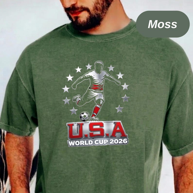 May include: A moss green short-sleeve t-shirt featuring a graphic of a soccer player kicking a ball surrounded by a circle of white stars. Below the player, the text reads U.S.A. WORLD CUP 2026 in bold red and white lettering on the chest.