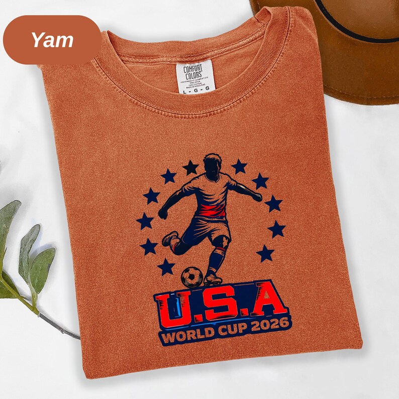May include: A rust-colored crew neck t-shirt featuring a graphic of a soccer player kicking a ball surrounded by a circle of dark blue stars. Below the player, the text reads U.S.A WORLD CUP 2026 in bold blue and red lettering on the front.