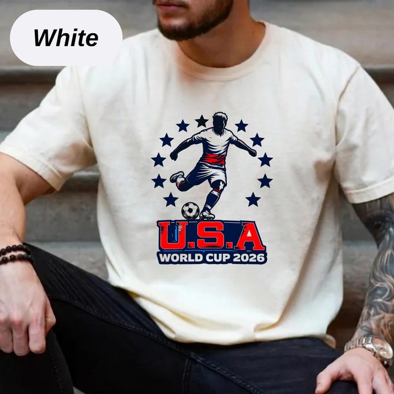 May include: A white short-sleeve t-shirt featuring a graphic of a soccer player kicking a ball, surrounded by a circle of blue stars. Below the player, the text reads U.S.A WORLD CUP 2026 in bold red and blue lettering on the front of the shirt.