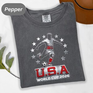 May include: A charcoal gray t-shirt featuring a graphic of a soccer player kicking a ball, surrounded by a circle of white stars. Below the player, the text reads U.S.A WORLD CUP 2026 in bold red and white lettering. The shirt has a crew neckline.