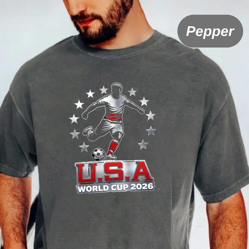 May include: A charcoal gray t-shirt featuring a graphic of a soccer player kicking a ball surrounded by a circle of white stars. Below the player, the text reads U.S.A WORLD CUP 2026 in bold red and white lettering. The shirt color is labeled Pepper.