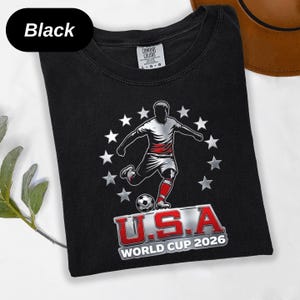 May include: A black t-shirt featuring a graphic of a soccer player kicking a ball, surrounded by a circle of white stars. Below the player, the text reads U.S.A WORLD CUP 2026 in red and white lettering. The shirt has a Comfort Colors brand label.