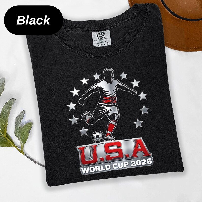 May include: A black t-shirt featuring a graphic of a soccer player kicking a ball, surrounded by a circle of white stars. Below the player, the text reads U.S.A WORLD CUP 2026 in red and white lettering. The shirt has a Comfort Colors brand label.