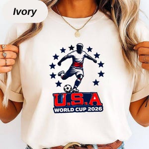 May include: An ivory t-shirt featuring a graphic of a soccer player kicking a ball, surrounded by a circle of dark blue stars. Below the player, the text reads U.S.A. WORLD CUP 2026 in bold red, white, and blue lettering on a dark blue background.