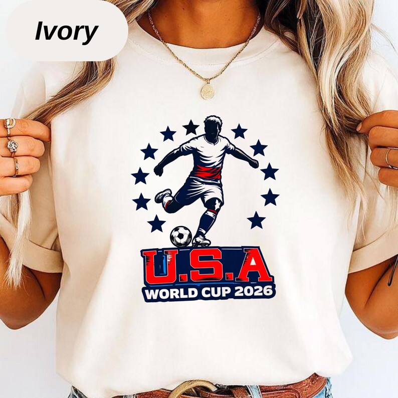 May include: An ivory t-shirt featuring a graphic of a soccer player kicking a ball, surrounded by a circle of dark blue stars. Below the player, the text reads U.S.A. WORLD CUP 2026 in bold red, white, and blue lettering on a dark blue background.