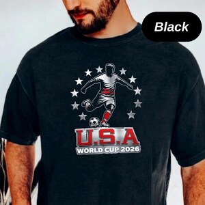 May include: A black t-shirt featuring a graphic of a soccer player kicking a ball, surrounded by a circle of white stars. Below the player, the text reads U.S.A. WORLD CUP 2026 in red and white lettering. The design is centered on the chest.