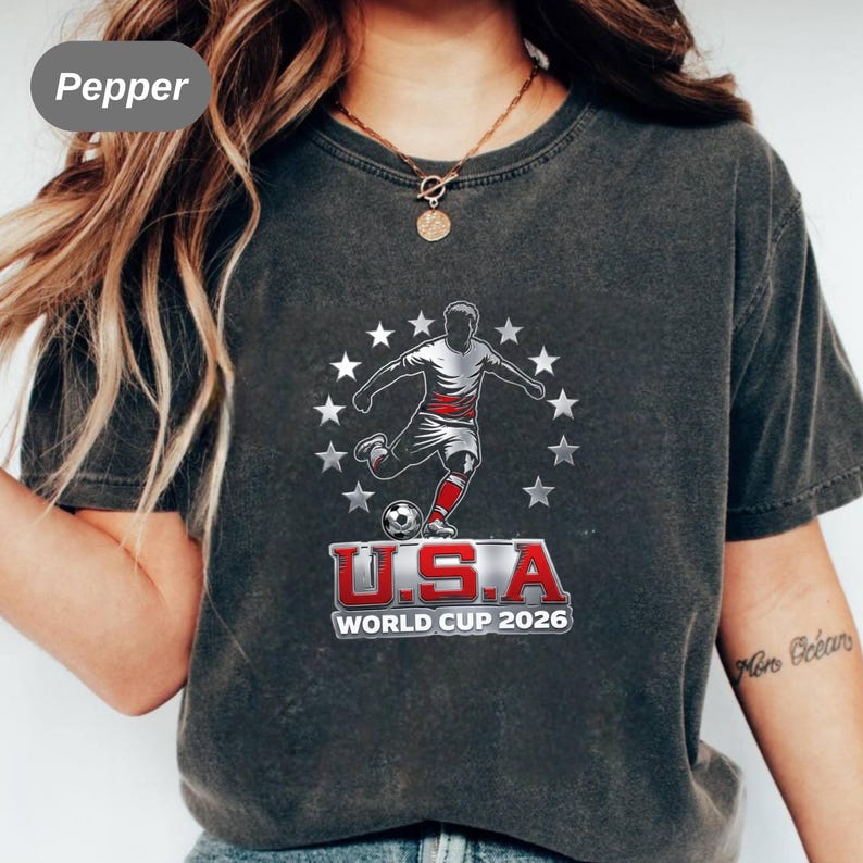 May include: A charcoal grey t-shirt featuring a graphic of a soccer player kicking a ball, surrounded by a circle of white stars. Below the player, the text reads U.S.A WORLD CUP 2026 in bold red and white lettering with a metallic silver outline.