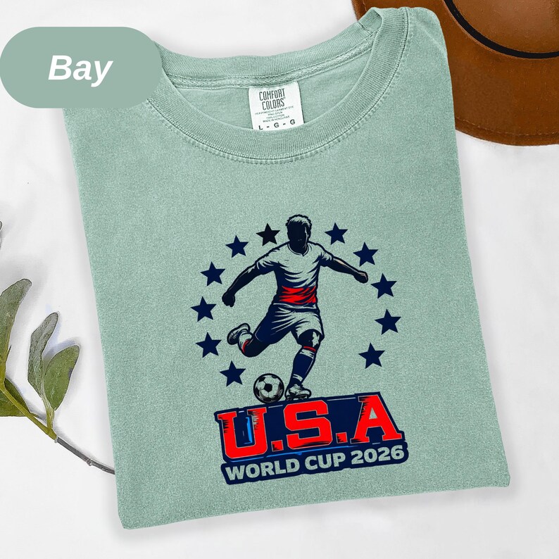 May include: A bay-colored t-shirt featuring a graphic of a soccer player kicking a ball, surrounded by a circle of dark blue stars. Below the player, the text reads U.S.A. WORLD CUP 2026 in bold red and blue lettering. The shirt has a Comfort Colors label.