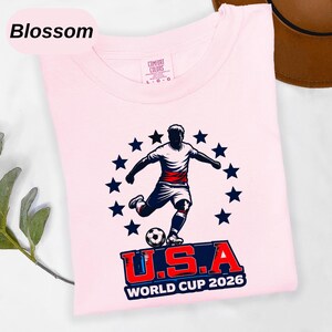 May include: A light pink t-shirt featuring a graphic of a soccer player kicking a ball surrounded by a circle of dark blue stars. Below the player, the text reads U.S.A WORLD CUP 2026 in bold red and blue lettering. The shirt has a Comfort Colors label.