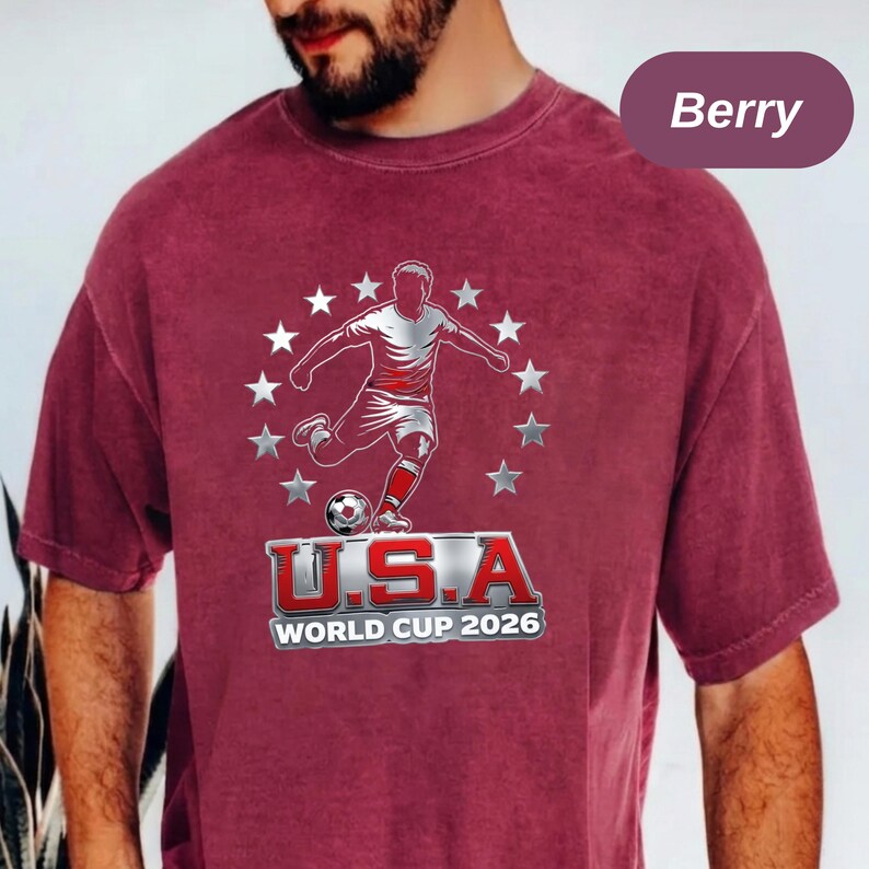 May include: A berry-colored short-sleeve t-shirt featuring a graphic of a soccer player kicking a ball surrounded by a circle of stars. Below the player, the text reads U.S.A WORLD CUP 2026 in bold, metallic-style lettering with a red outline.
