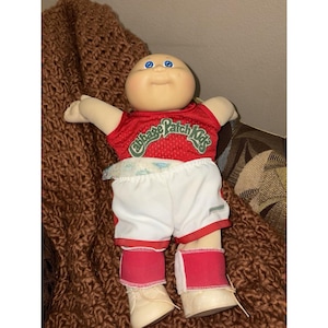 May include: A Cabbage Patch Kids doll wearing a red jersey with the words "Cabbage Patch Kids" in green, white shorts, and red leg warmers. The doll has blue eyes and a smiling expression.