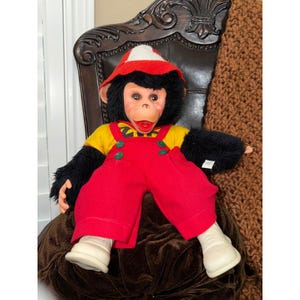 May include: A vintage monkey doll with a red and white hat, black fur, and a yellow shirt under red overalls. The doll has a painted face, white shoes, and is seated on a brown velvet cushion.