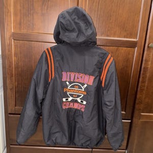 May include: Black hooded jacket with orange stripes on the sleeves. The back of the jacket features the text "DIVISION CHAMPS" with hockey sticks and the team name "Phantoms" in pink and white.