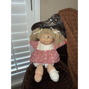 May include: A vintage Cabbage Patch Kids doll with blonde yarn pigtails and green eyes. The doll wears a red and white checkered dress with a white collar and white lace-trimmed bloomers. A white crocheted shoe is on one foot.