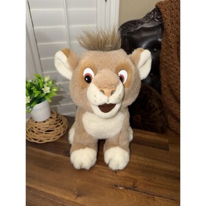 May include: A plush lion cub toy with a light brown body, cream-colored belly and paws, and a tuft of brown hair. The toy has large, expressive eyes with red accents and a smiling mouth. A cute stuffed animal.