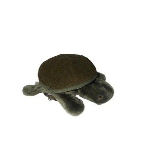 May include: A plush turtle toy in shades of green. The turtle has a textured, dark green shell and lighter green, fuzzy body, with small, beady eyes. The toy is lying on a white background.
