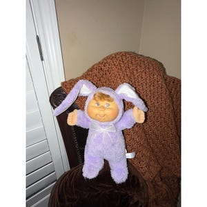 May include: A Cabbage Patch Kids doll with curly light brown hair and a round face, wearing a fuzzy light purple hooded onesie with long bunny ears attached to the hood and a small white ribbon bow centered on the chest area of the garment.