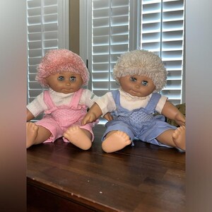 May include: Two vintage baby dolls with blue eyes and soft, fluffy hair. One doll wears pink overalls, the other wears blue overalls. Both dolls have a white shirt underneath and are sitting on a wooden surface.