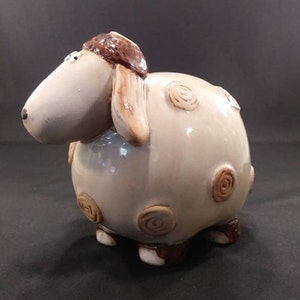May include: A ceramic sheep figurine with brown spots and a brown hat. The sheep is light gray and has a glossy finish.