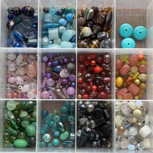 May include: A clear plastic organizer filled with various beads in different colors and shapes. Beads include blue, green, red, purple, and clear, with round, oval, and irregular shapes. The beads are stored in individual compartments.