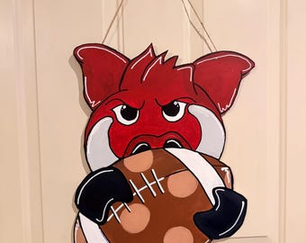 May include: A wooden door hanger featuring a red and white cartoon hog holding a brown football with white laces. The hog has an angry expression. The door hanger is suspended by a natural twine string.