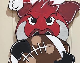 May include: A wooden cutout of a red Razorback hog wearing a Santa hat, holding a brown football with white laces. The hog has an angry expression. The cutout is painted with black and white accents.