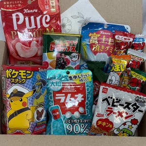 Puede incluir: Una caja de cartón llena de varios bocadillos y caramelos japoneses. La caja contiene paquetes coloridos de gominolas, caramelos y bocadillos, incluyendo una bolsa roja de caramelos con sabor a melocotón, una lata de caramelos de frutas y ramen.