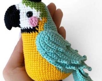 May include: A handmade crochet parrot in yellow, blue, white, and green. The bird has a black beak, a small black hat, and a pink cheek. The parrot is a whimsical, handcrafted item.