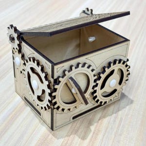May include: A wooden box with a hinged lid and decorative gear-shaped accents. The box is made of light-colored wood and features three gear wheels on the front and side. The lid is slightly ajar, revealing the box's interior.