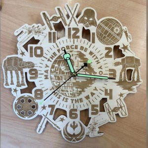 Puede incluir: Un reloj de pared circular de madera con imágenes de Star Wars cortadas con láser, incluyendo a Darth Vader, la Estrella de la Muerte, caminantes AT-AT y cazas estelares. La esfera muestra el texto MAY THE FORCE BE WITH YOU THIS IS THE WAY.