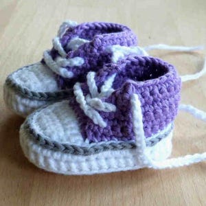 Peut inclure: Une paire de chaussons de bébé crochetés à la main. Les chaussures sont blanches avec des accents violets et une bordure grise. Des lacets blancs sont noués en nœud. Les chaussures ressemblent à des baskets miniatures.