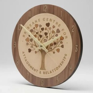 Könnte beinhalten: Eine runde Wanduhr aus Holz mit einem dunkelbraunen Außenring und hellen Ziffern von 1 bis 12. Die Mitte besteht aus einer helleren Holzeinlage, die einen Baum mit herzförmigen Blättern und den Text BRISBANE CENTRE FOR ATTACHMENT & RELATIONSHIPS zeigt.