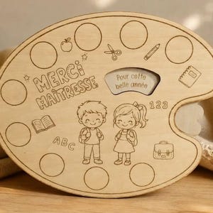 May include: A wooden palette-shaped gift for a teacher, engraved with the words "Merci Maîtresse" and "Pour cette belle année". It features illustrations of school supplies, children, and the numbers "123" and "ABC".
