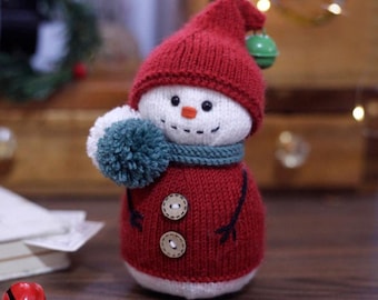May include: A handmade snowman figure with a red knit hat and sweater, a white face, and a teal scarf and pom-pom. The snowman has a small orange nose, black stitched mouth, and two wooden buttons on the sweater. A green bell hangs from the hat.