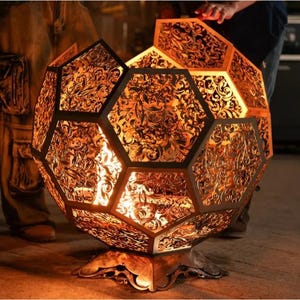 Puede incluir: Un brasero decorativo esférico con un diseño geométrico. El brasero de metal presenta intrincados recortes florales, que permiten que el cálido resplandor del fuego brille. Se asienta sobre una base decorativa y está rodeado de personas.