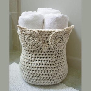 Peut inclure: Un panier en forme de hibou, crocheté en crème, contenant des serviettes blanches roulées. Le panier présente de grands détails circulaires pour les yeux et un bec pointu. Les serviettes sont soigneusement disposées à l'intérieur.