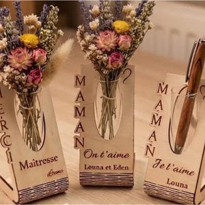 Op de afbeelding: Drie houten bureau-organizers met bloemstukken en een pen. De organizers hebben uitgesneden tekst met de woorden "Merci", "Maman" en "Je t'aime". Elke organizer heeft een glazen vaas of penhouder.