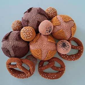 Op de afbeelding: Een verzameling handgemaakte gehaakte voedselitems, waaronder ronde broodjes in bruin- en beigetinten en bruine pretzels met witte details. De items zijn gerangschikt op een lichtblauwe ondergrond.